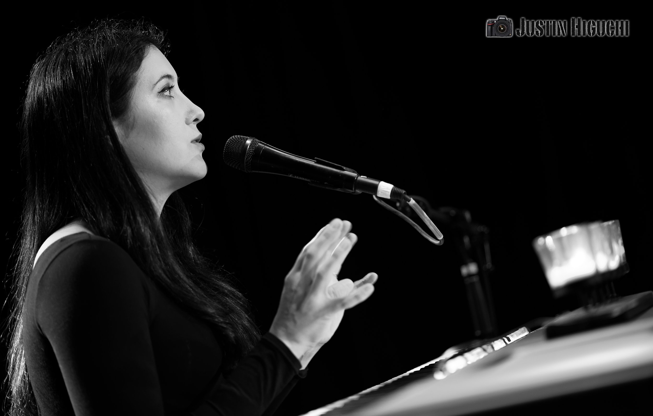 A black & white photo of Carlton, sitting at a piano, speaking into a microphone with her hands up.
